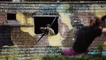 A person aiming a paintball gun through a window of a building with peeling paint and graffiti.