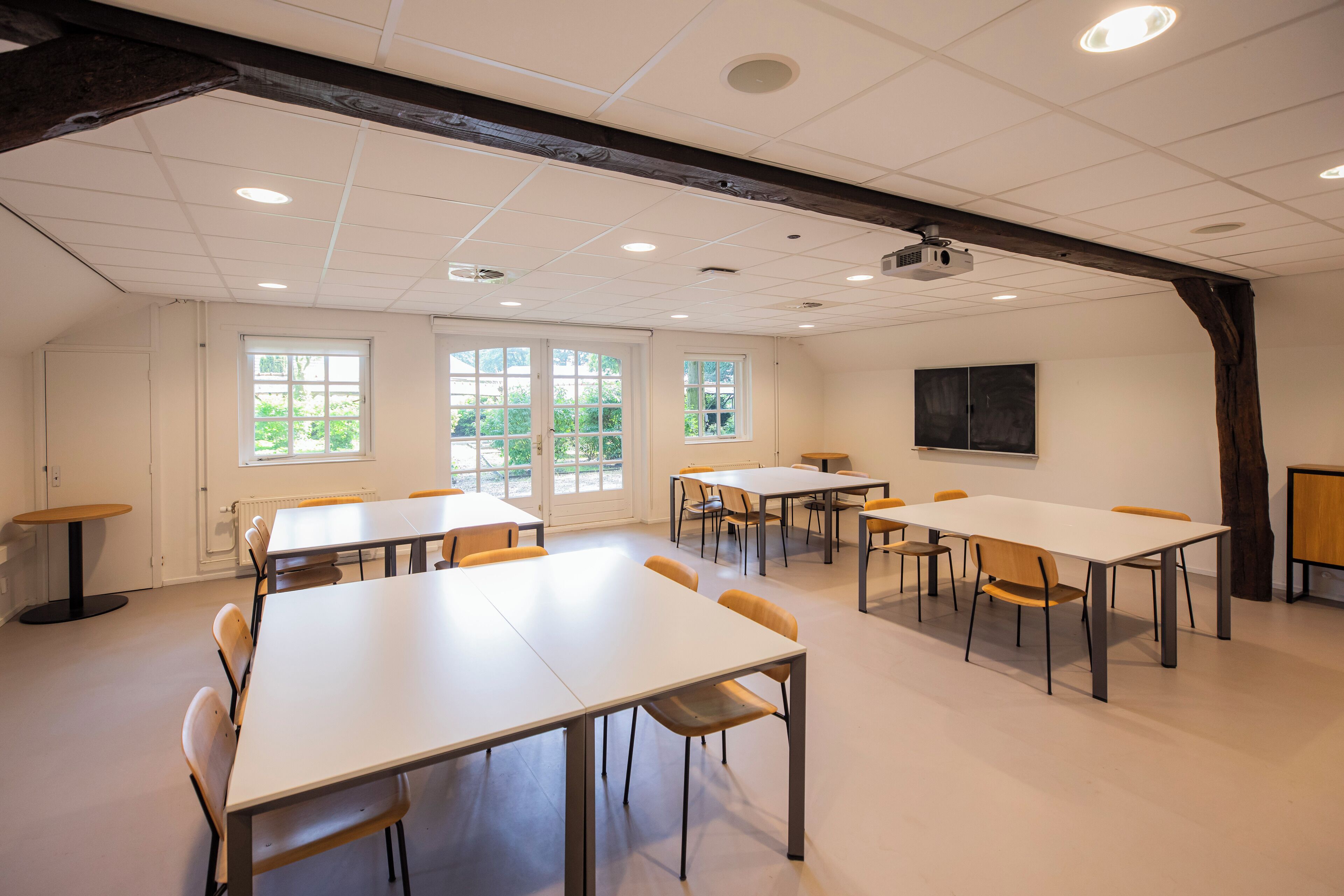 A conference room with multiple tables and chairs, large windows, and a projector screen on the wall.