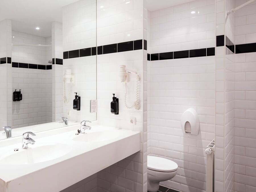 A modern bathroom with white tiles, a large mirror, two sinks, and a toilet.