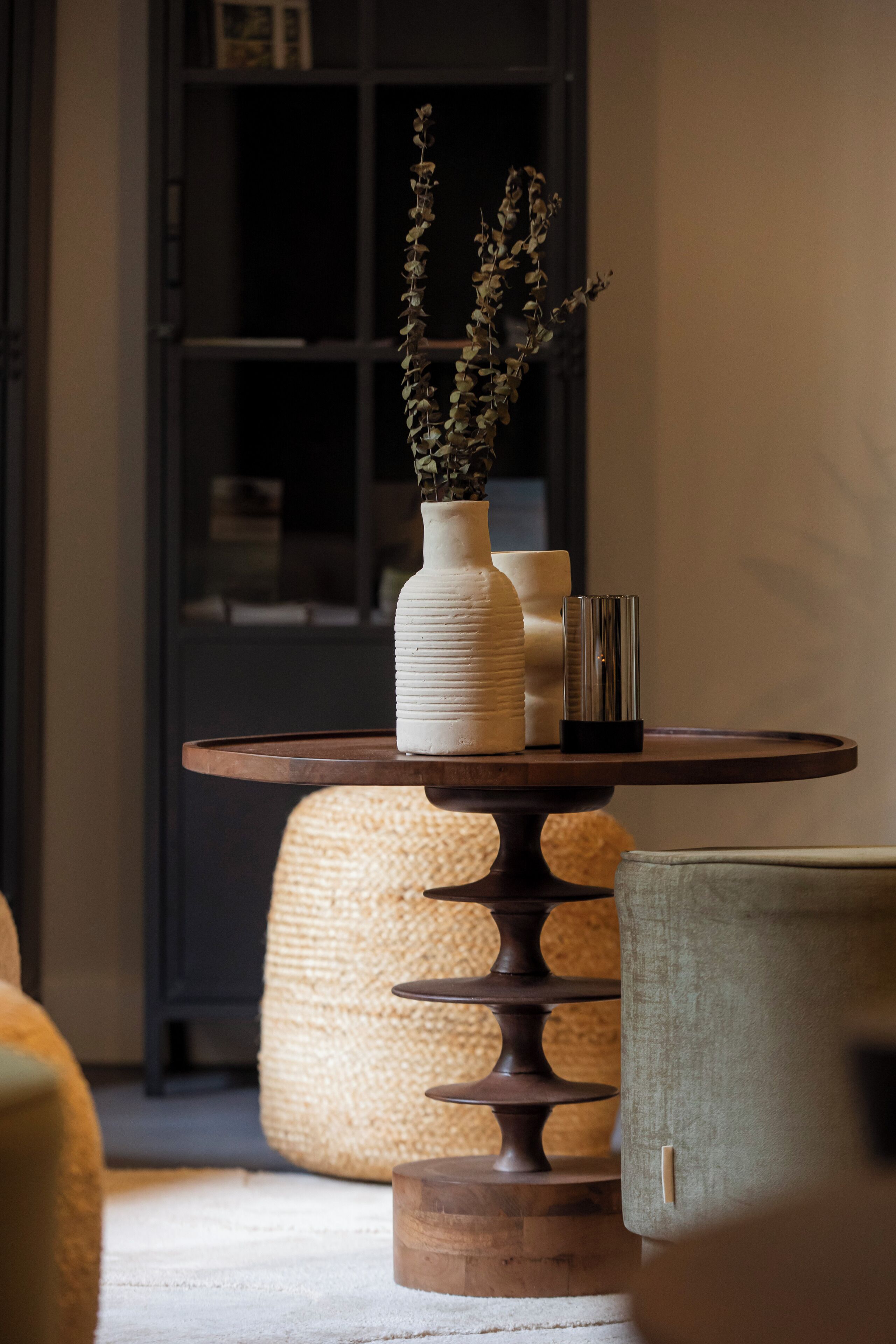 Una mesa redonda de madera con un jarrón blanco y velas, un pouf tejido y una silla tapizada de verde.