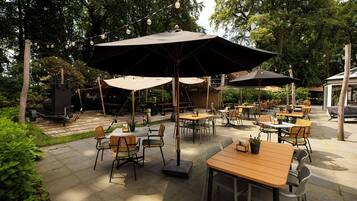 Een terras buiten met meerdere tafels en stoelen, omgeven door groen en bomen.