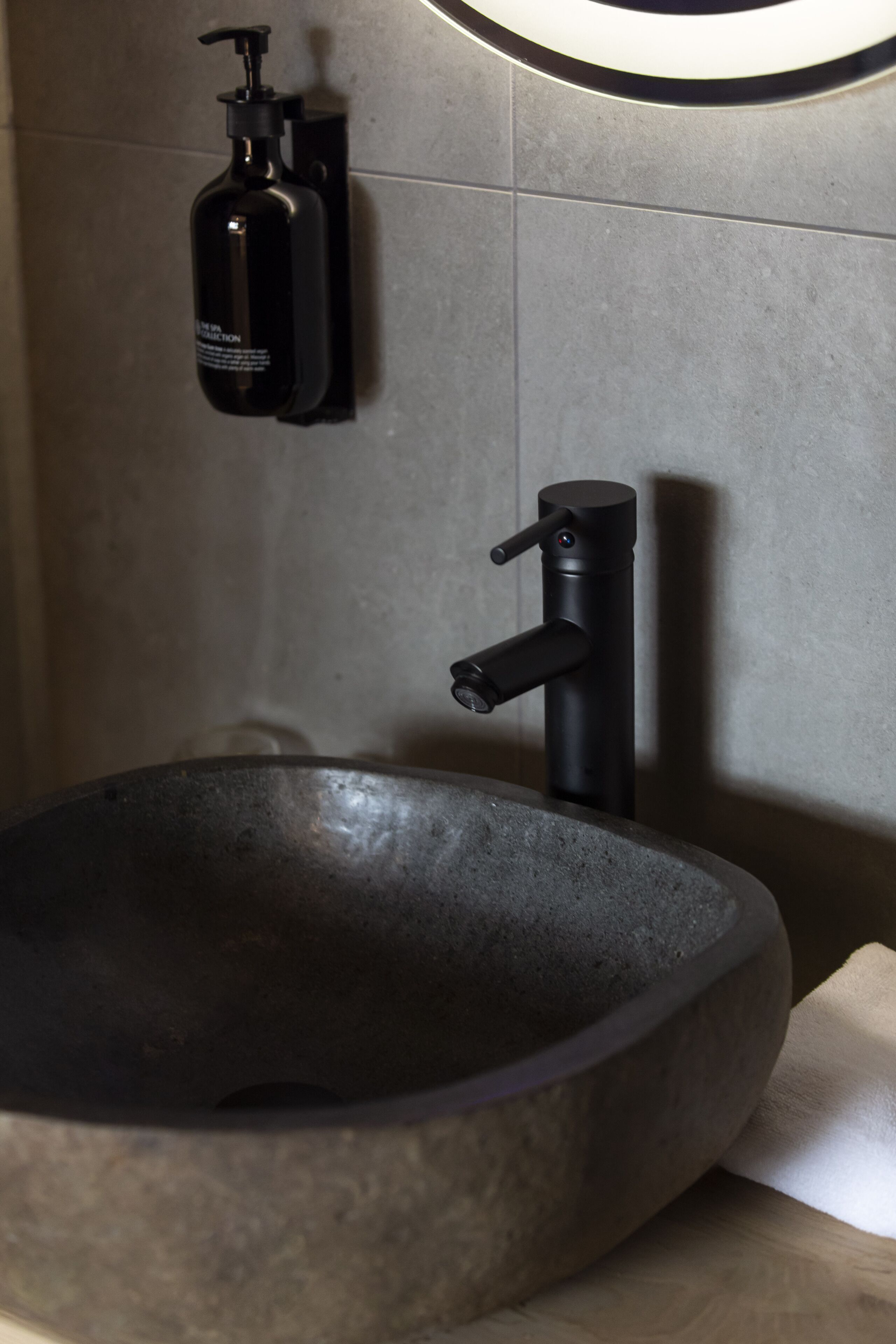 Ein modernes Badezimmer mit einem großen, runden Steinsink, einem schwarzen Wasserhahn und einer schwarzen Seifenspenderleiste an der Wand.