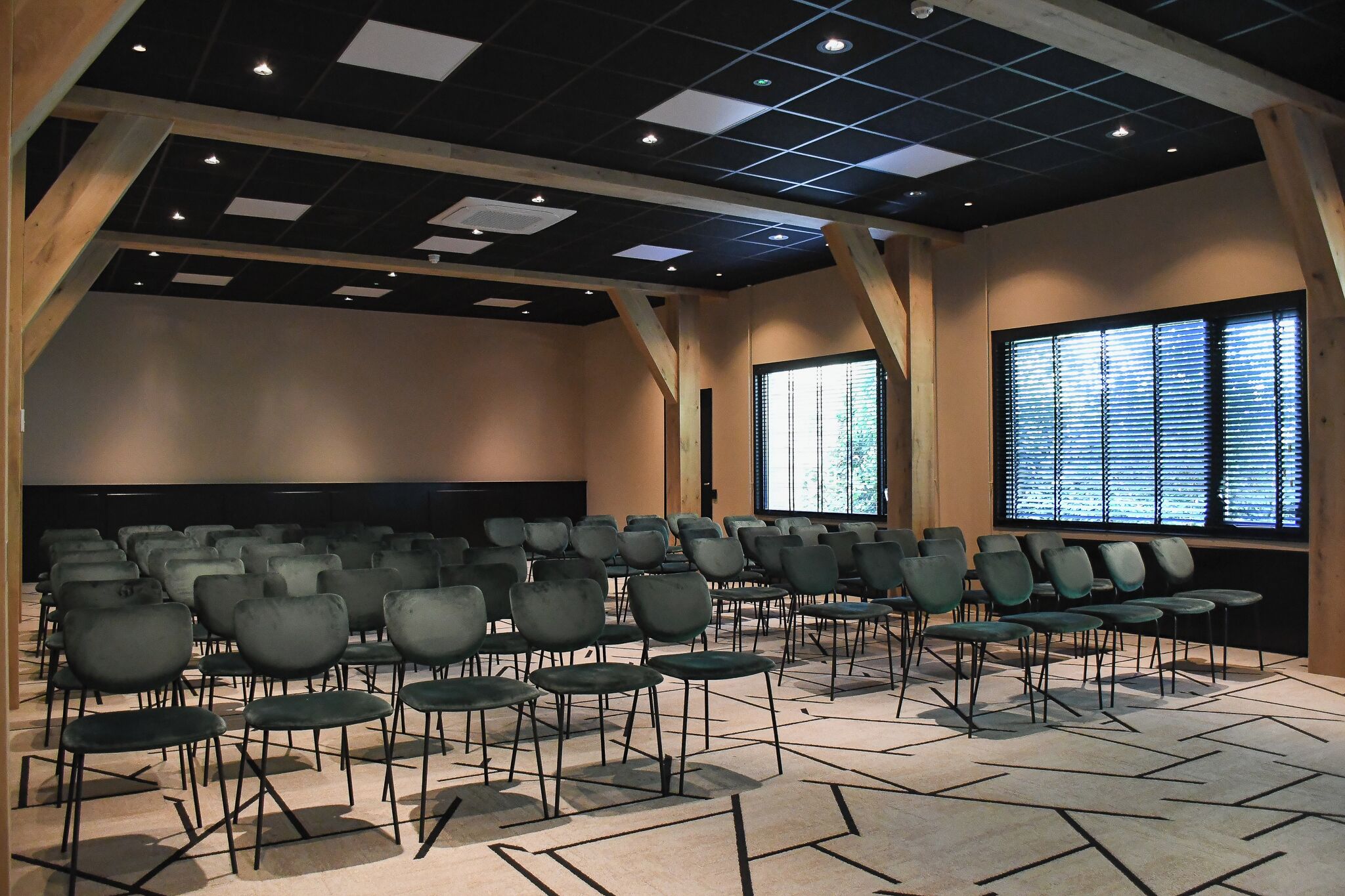 Un salón de conferencias con filas de sillas, una pantalla para proyector y un amplio ventanal con persianas.
