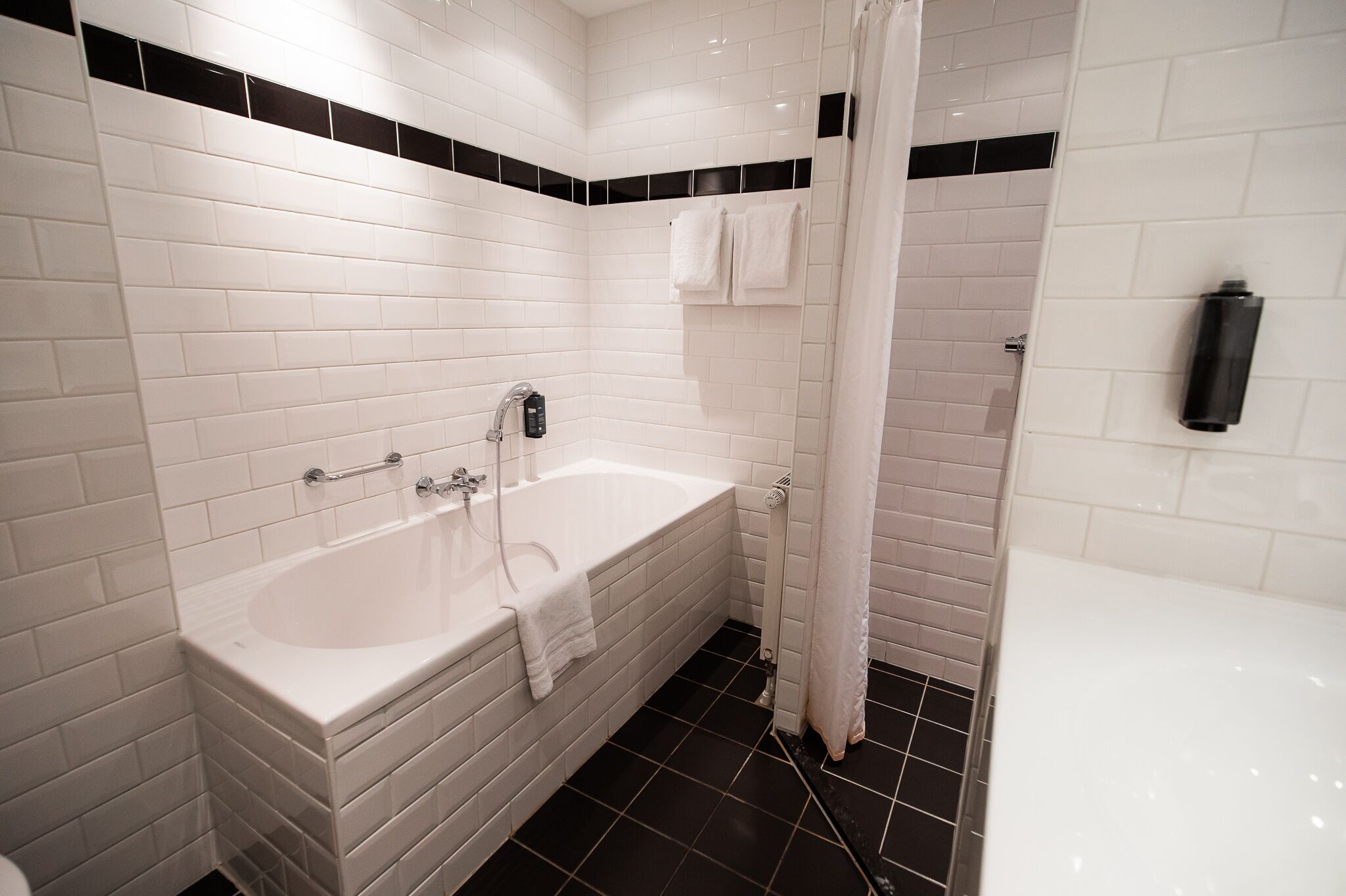 Un baño moderno con azulejos blancos tipo metro, una bañera blanca, zona de ducha y suelo negro.