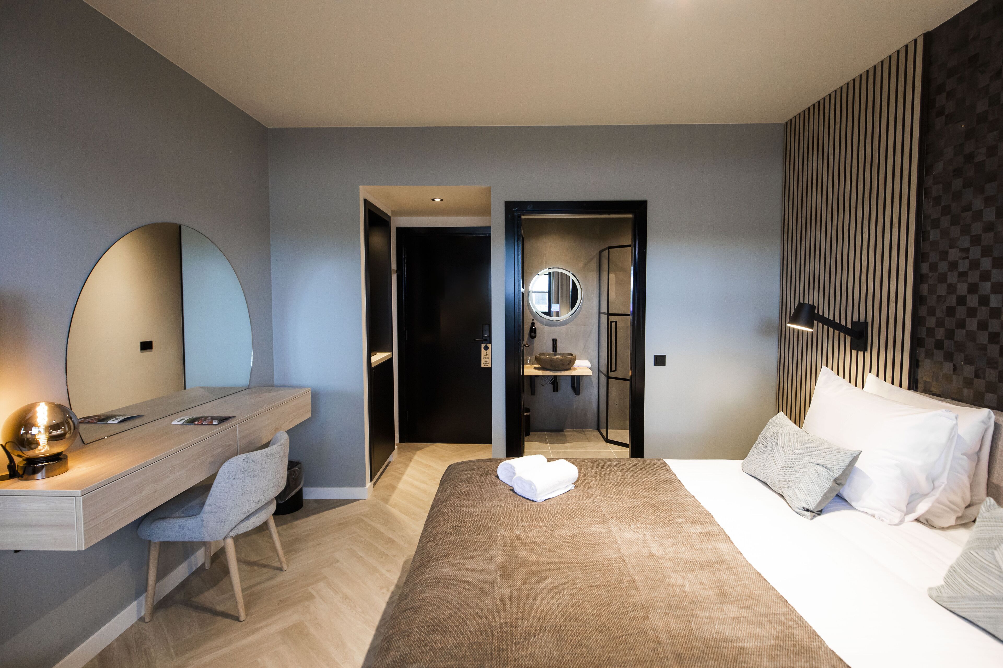 Et moderne hotelværelse med et træskrivebord, en stor seng med hvide og mønstrede puder, et rundt spejl og en ternet vægpanel.