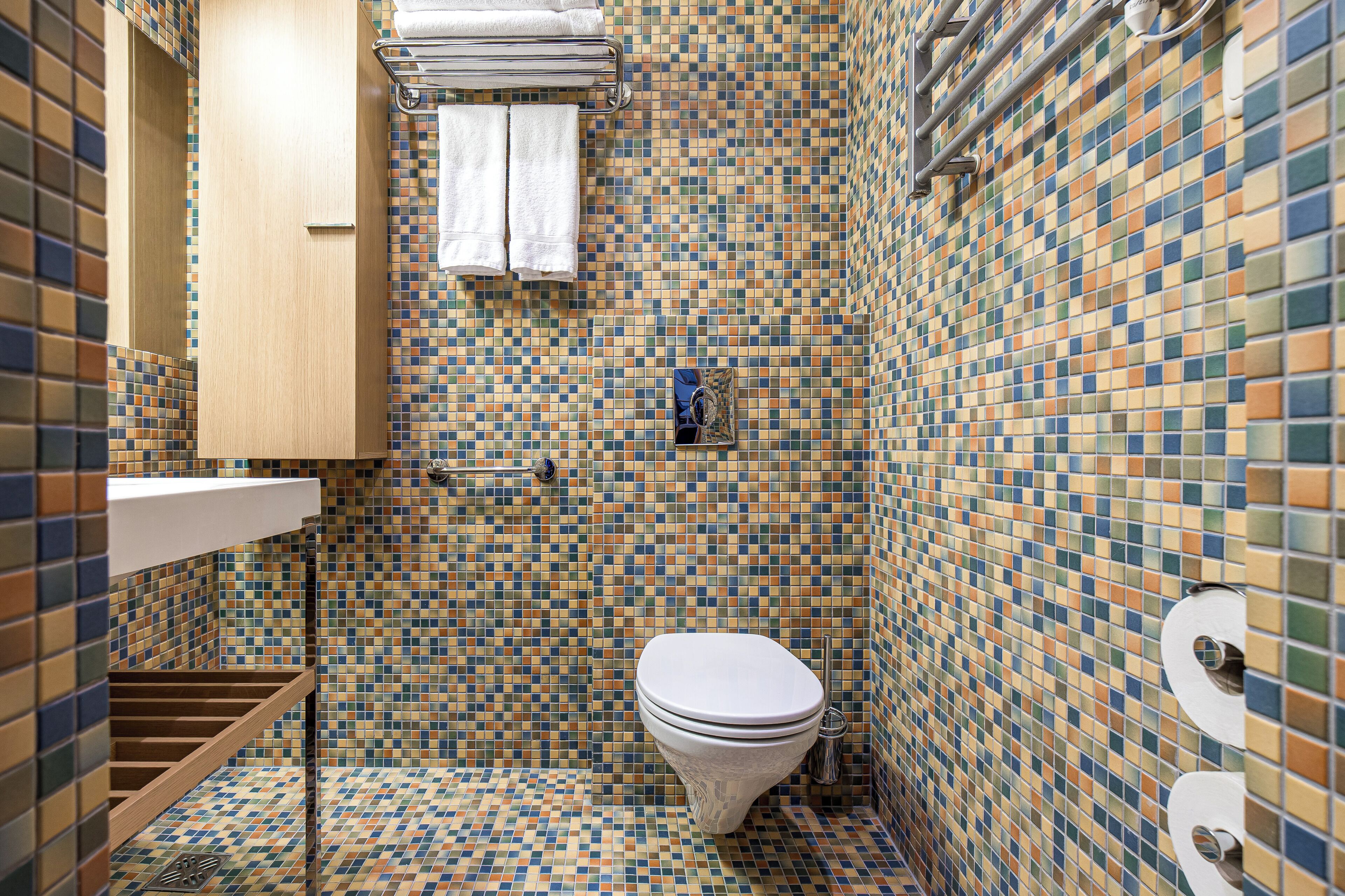 A bathroom with a mosaic tile wall, a toilet, a towel rack, and a wooden shelf.