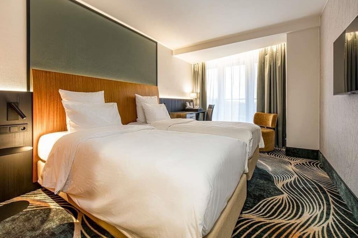 A hotel room with two beds, a desk, a chair, and a large window with curtains.