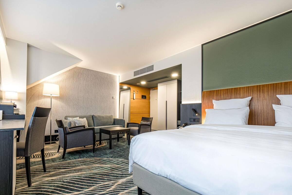 A hotel room with a bed, a desk, a sofa, a chair, a lamp, and a patterned carpet.