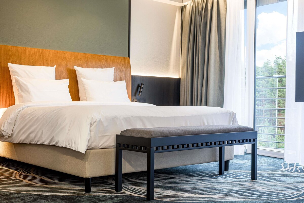 A modern hotel room with a large bed, a bench, and a view of greenery through the window.