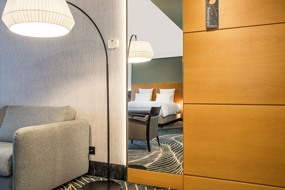 A hotel room with a sofa, a chair, a bed with white linens, a wooden wardrobe, and a mirror reflecting the room.