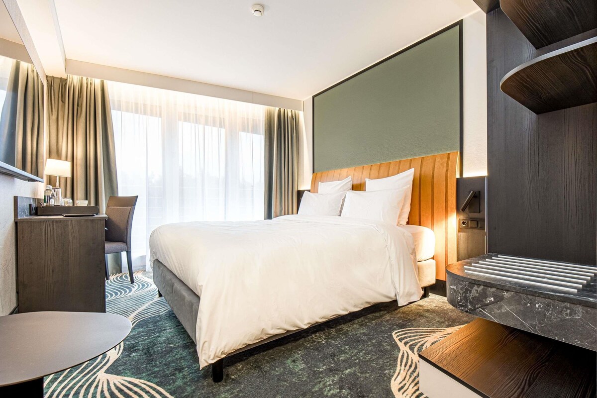 A modern hotel room with a large bed, a desk, a chair, and a view of the outdoors through sheer curtains.