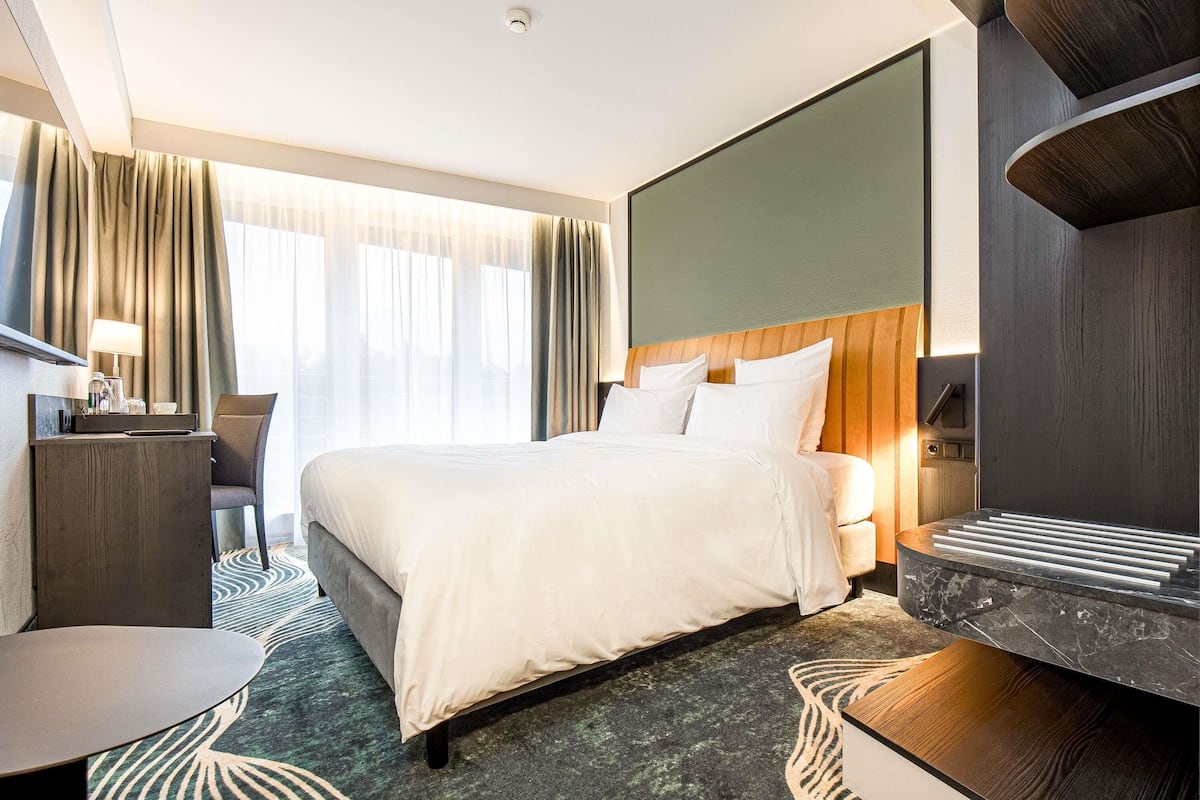 A modern hotel room with a large bed, a desk, a chair, and a view of the outdoors through sheer curtains.