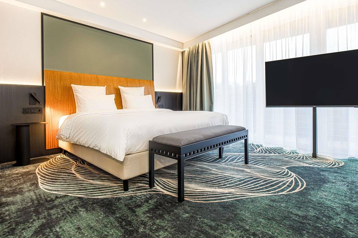 A modern hotel room with a large bed, a bench, a flat-screen TV, and a unique patterned carpet.