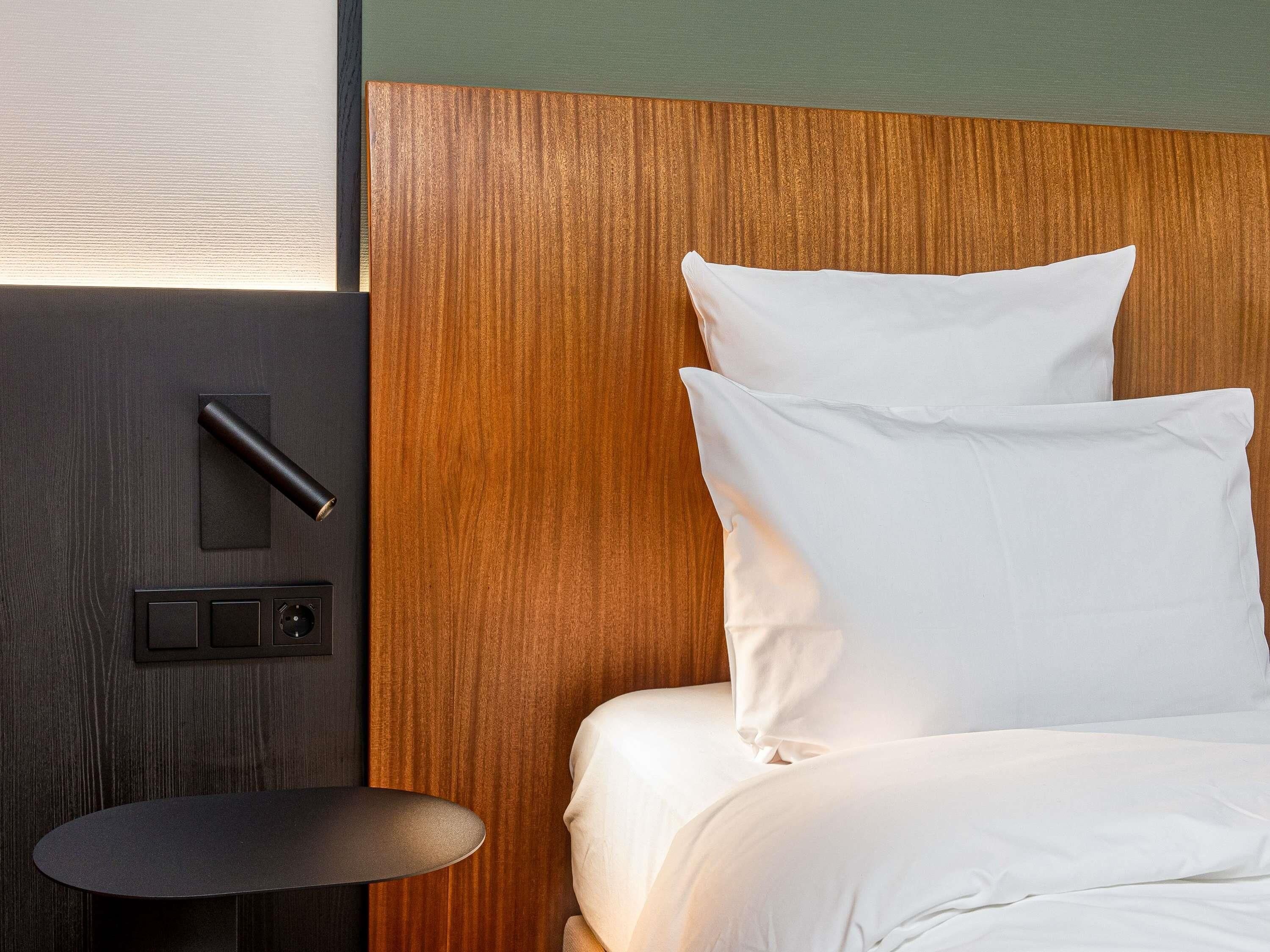 Ein Hotelzimmer mit Holz-Kopfteil, einem Bett mit weißer Bettwäsche, einem kleinen runden Tisch und einer an der Wand montierten Steuerung.