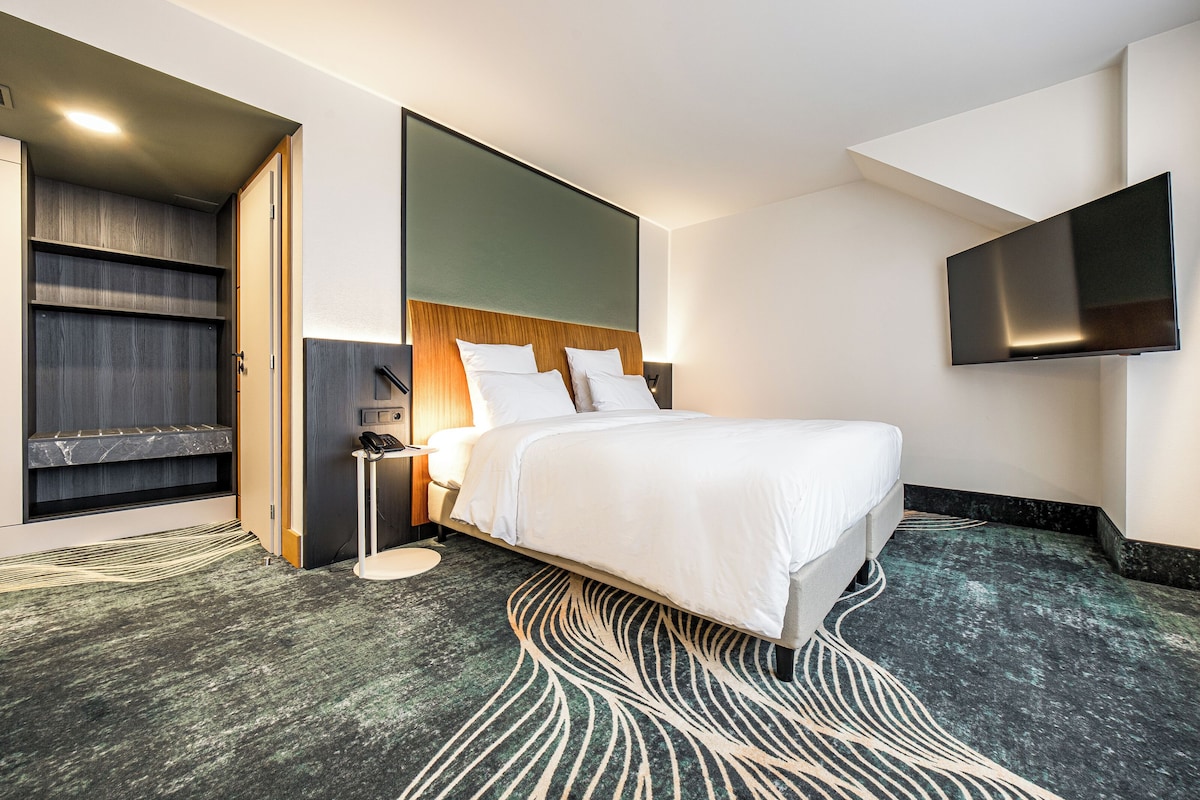 A modern hotel room with a large bed, a flat-screen TV mounted on the wall, a built-in bench with storage, and a patterned rug.