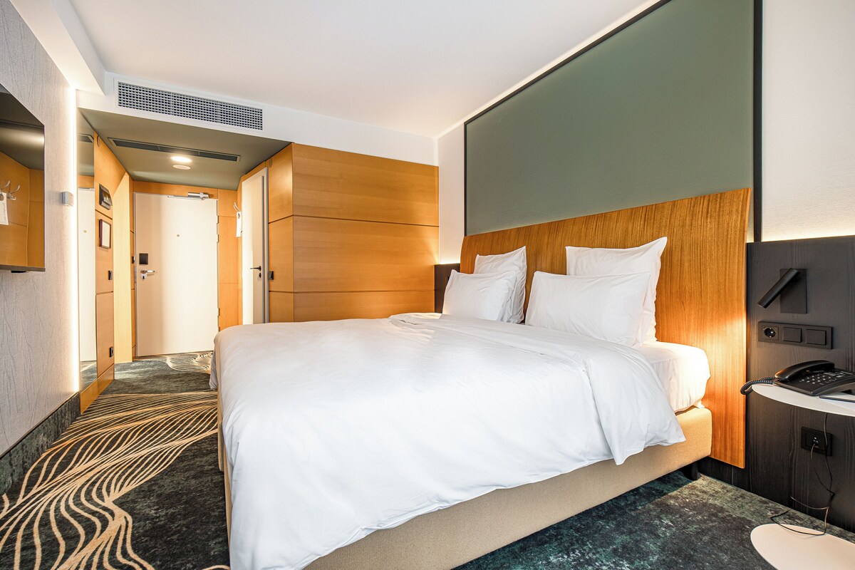 A modern hotel room with a large bed, a bedside table with a phone, a headboard, and a carpeted floor.
