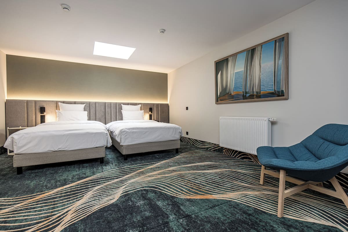 A hotel room with two beds, a blue armchair, a painting on the wall, and a patterned carpet.