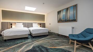 A hotel room with two beds, a blue armchair, a painting on the wall, and a patterned carpet.