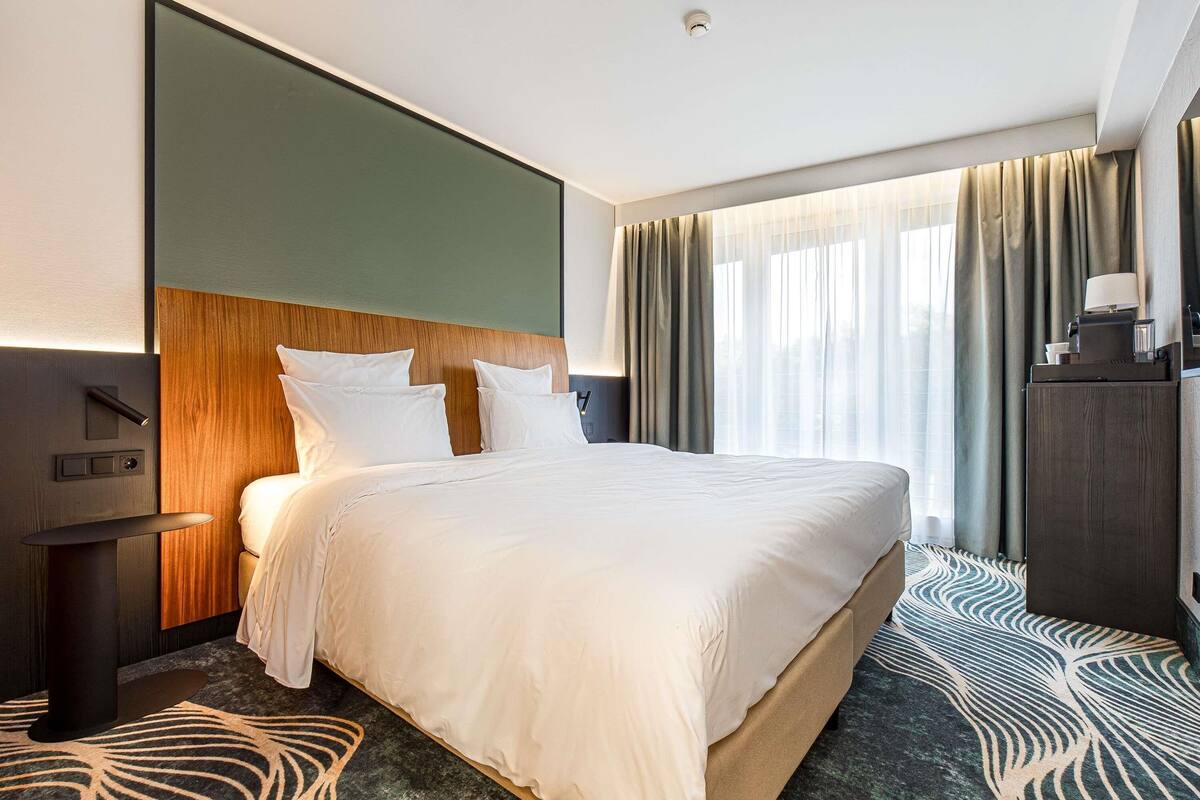 A hotel room with a large bed, a nightstand, a desk, and a window with curtains.