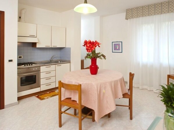 Una cocina funcional con armarios blancos, una estufa y un lavamanos. Una mesa de comedor con un florero rojo y una silla se encuentra en el centro.