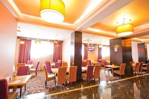A hotel lobby with multiple dining tables and chairs, a television screen, and a patterned carpet.
