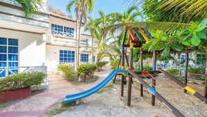 A playground with a slide and climbing structures, surrounded by palm trees and a building with large windows. - Hotel Tropical Inn (Cartagena)
