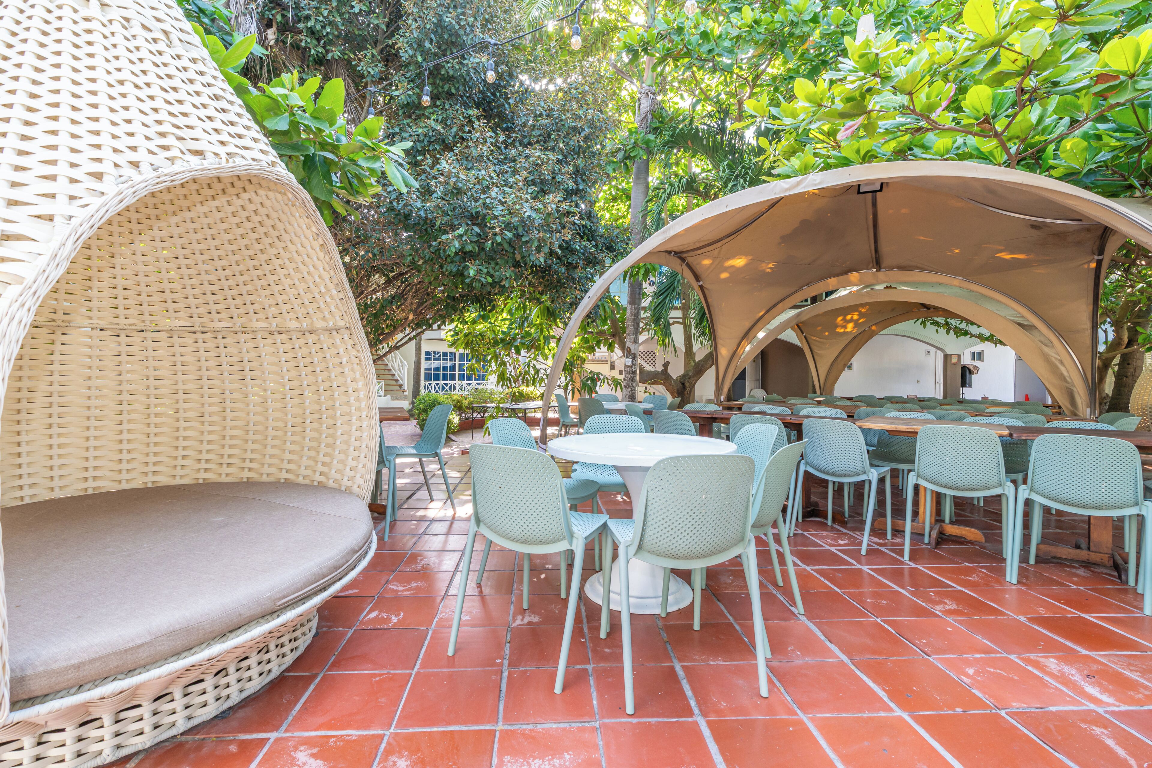 An outdoor dining area with wicker seating and tables, surrounded by greenery.