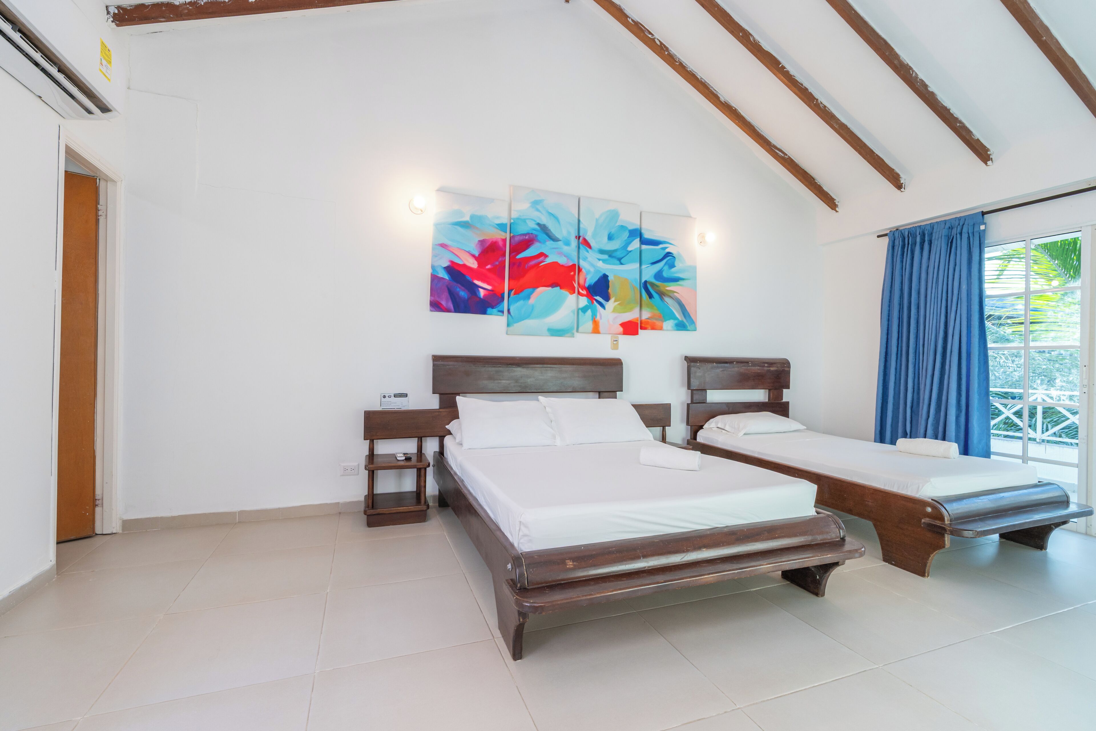 A room with two wooden beds, a large abstract painting, and a window with blue curtains.