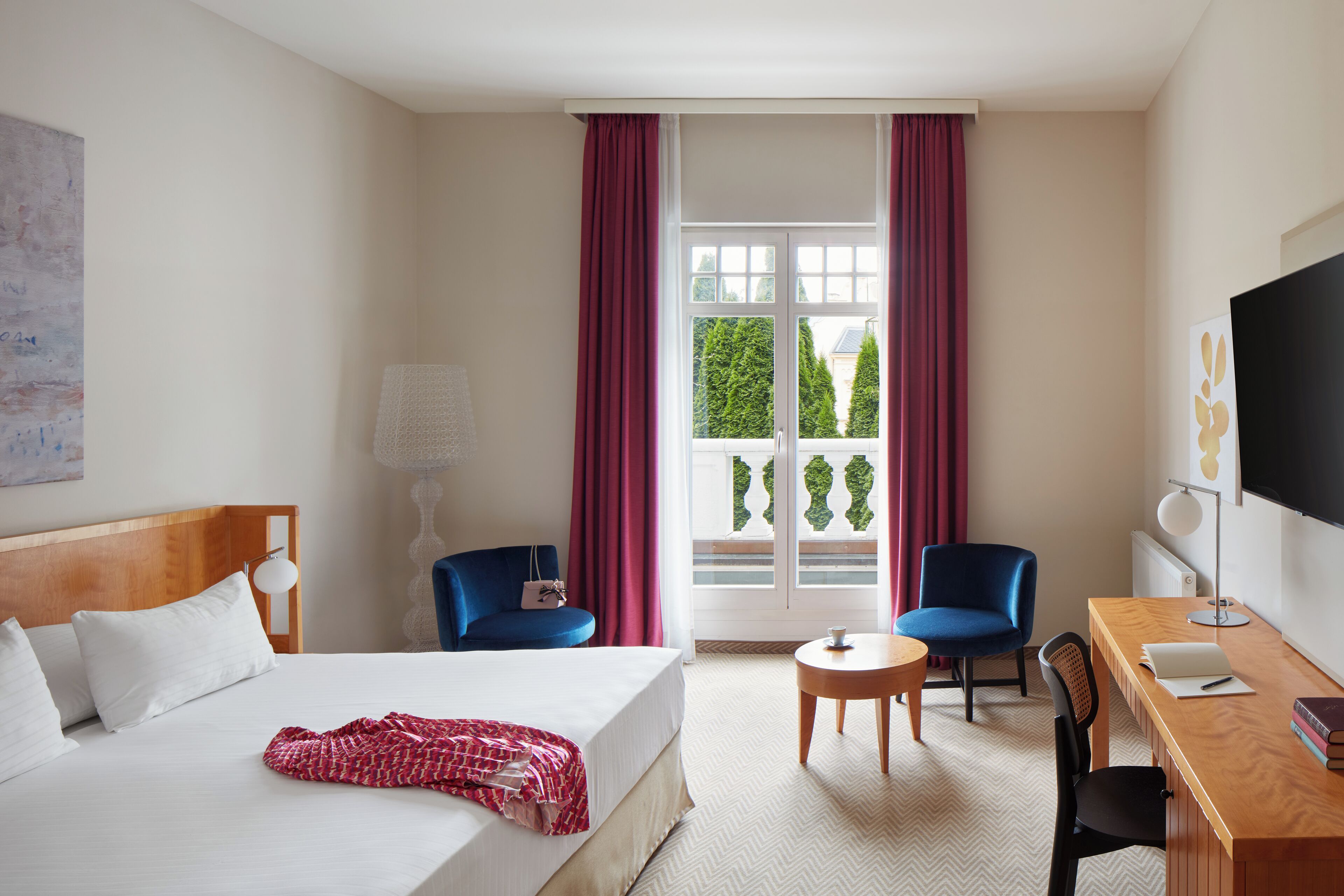 A hotel room with a bed, two chairs, a desk, a TV, and a window with curtains.