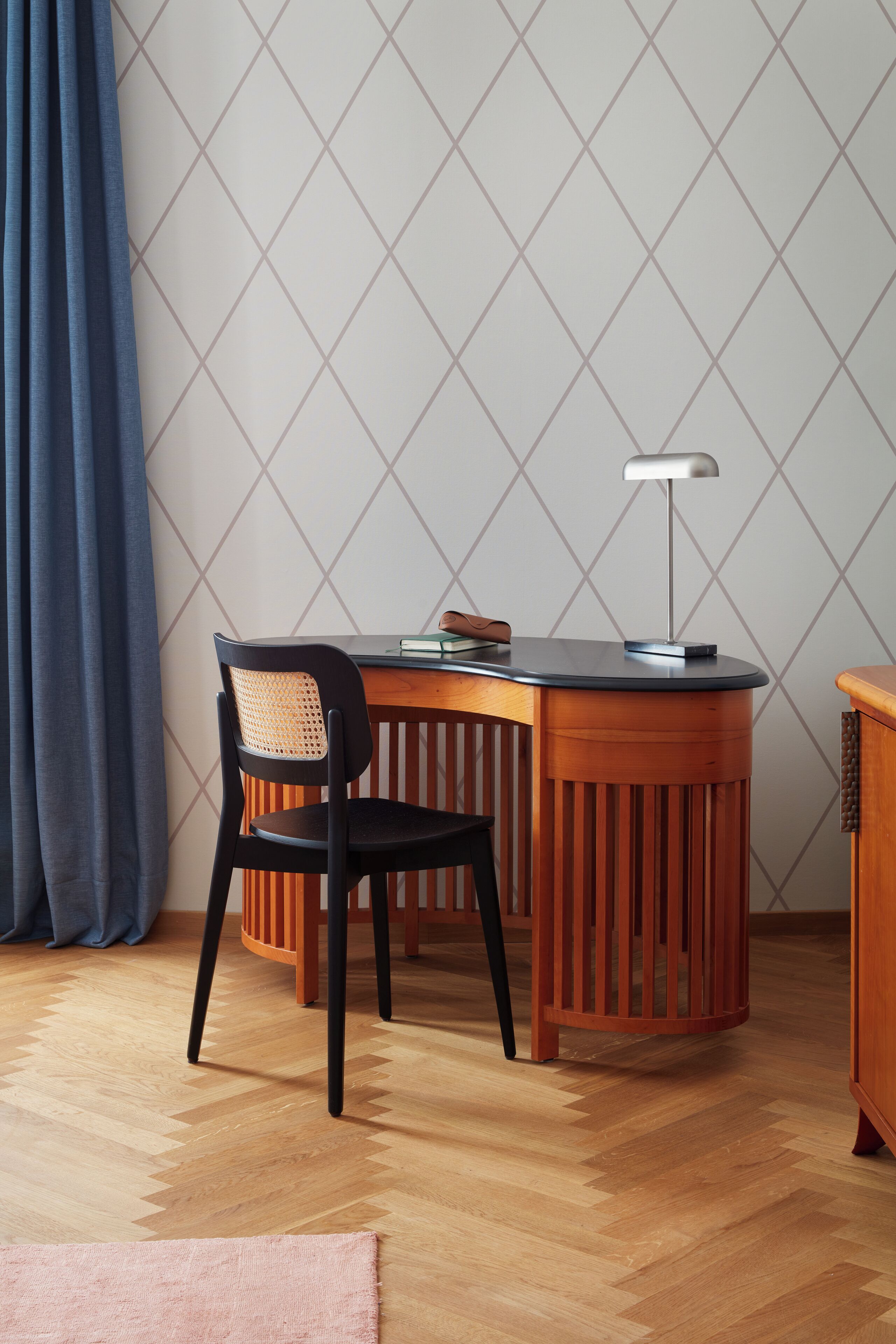 A corner room with a wooden desk, a black chair, a lamp, and a patterned wall.