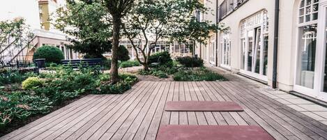 Una terraza de madera con dos esterillas de yoga, rodeada de vegetación y edificios.