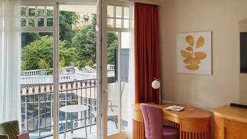 A hotel room with a bed, a desk, a chair, a table, and a balcony with a view of the city.