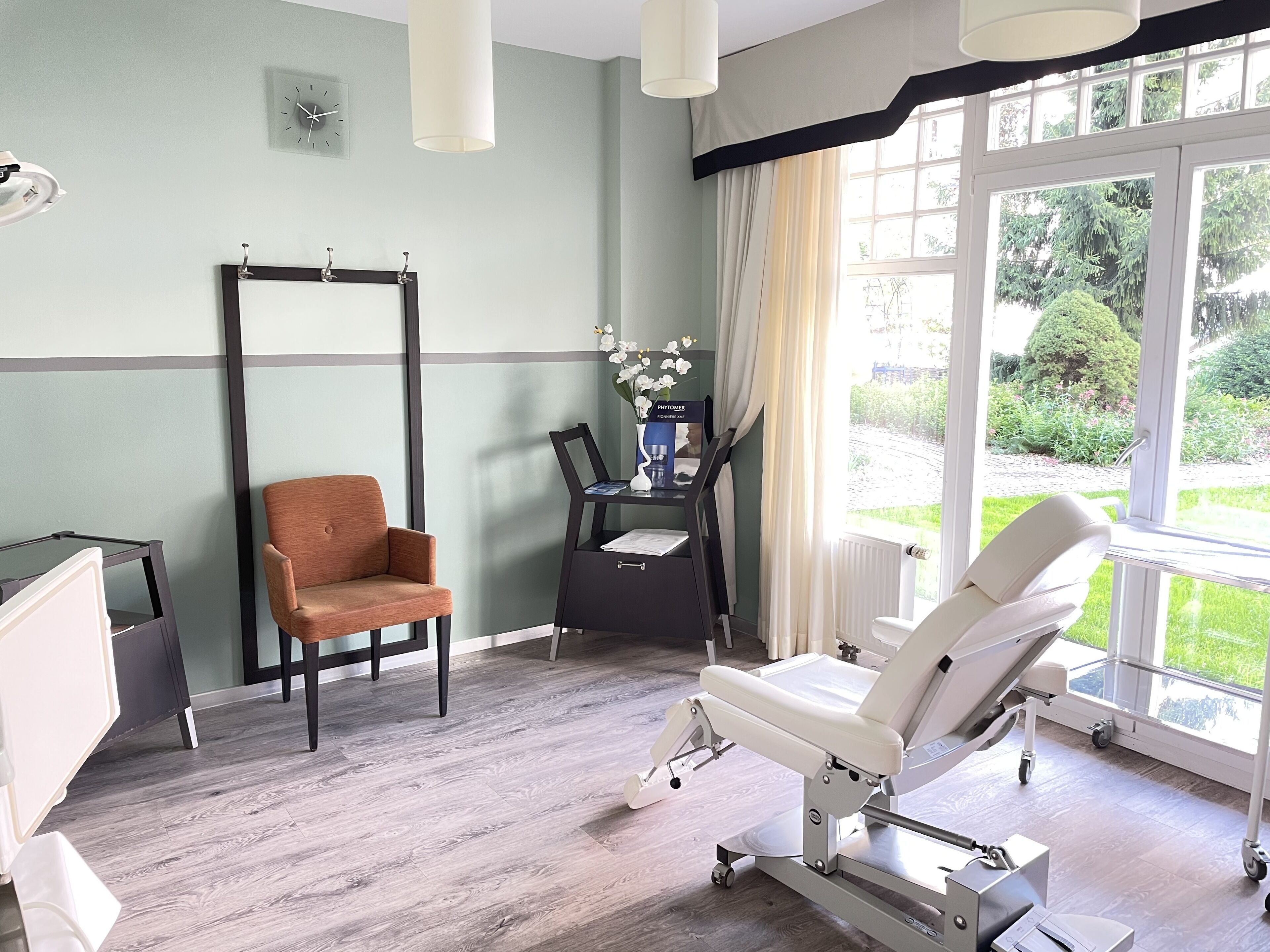 A clinical room with a patient bed, a chair, a small table with a vase of flowers, and a window with a view of greenery.
