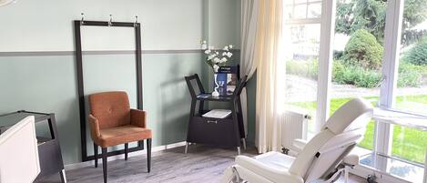 A clinical room with a patient bed, a chair, a small table with a vase of flowers, and a window with a view of greenery.