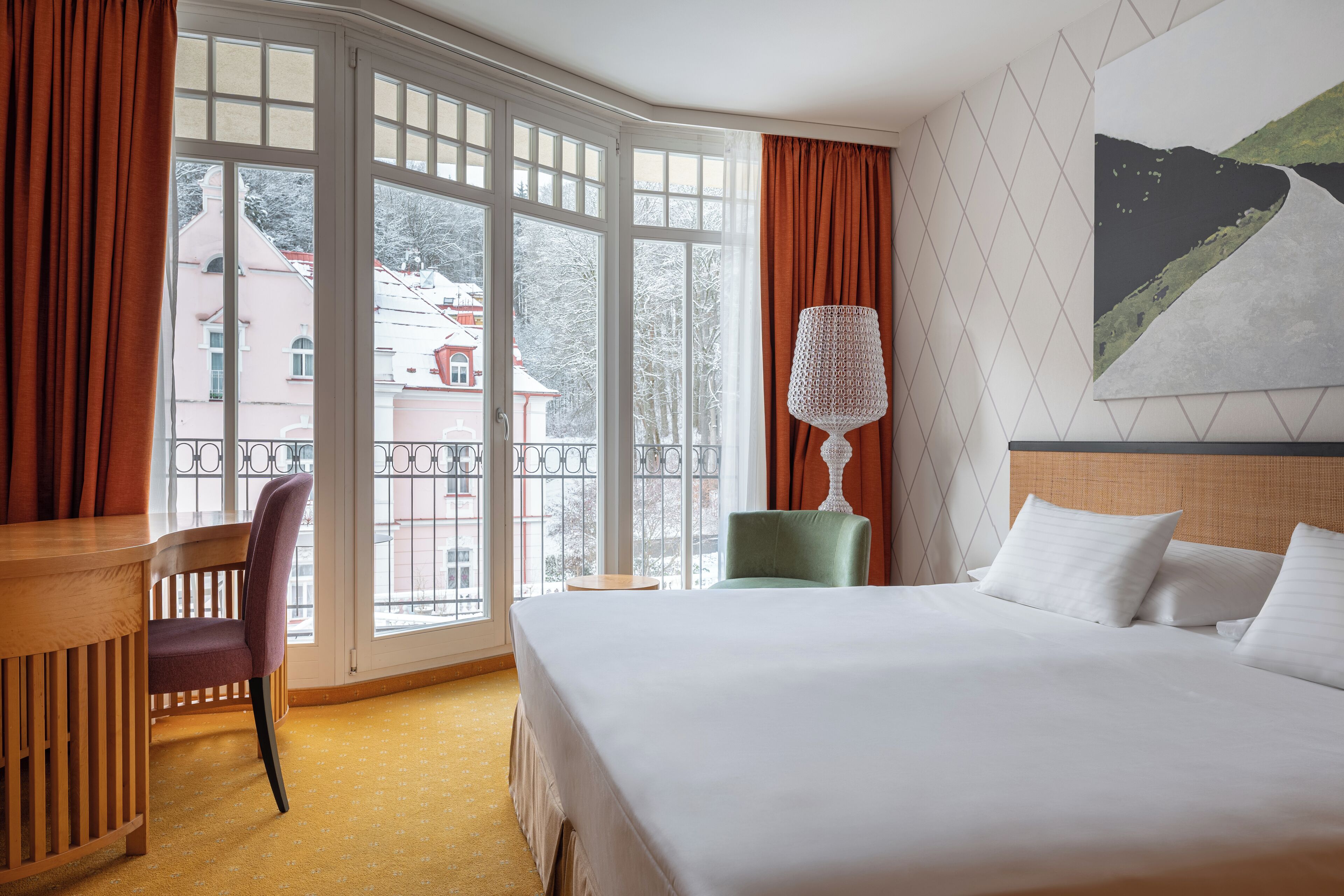 A hotel room with a large window, a bed with white linens, a green chair, a wooden side table, and a wall-mounted lamp.