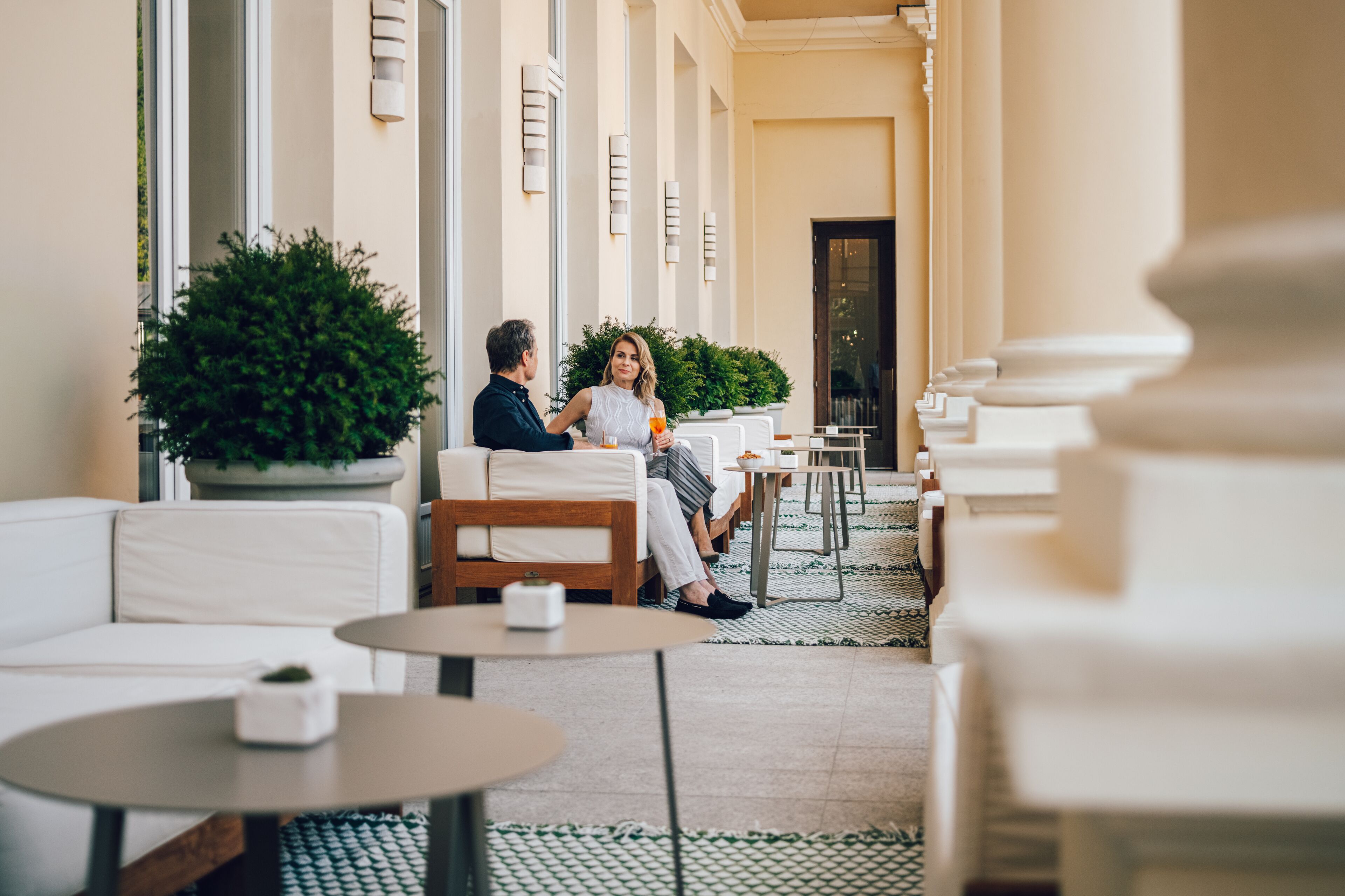 Una pareja sentada en un café con terraza, plantas en macetas y piso de baldosas.