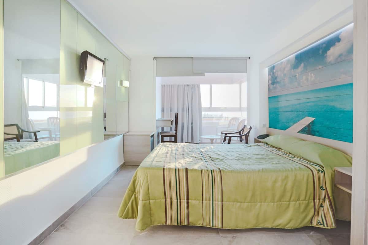 A hotel room with a bed, a TV mounted on the wall, a desk with a chair, and a large window.