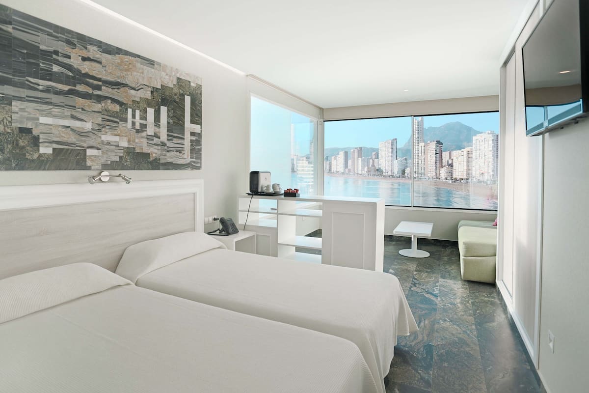 A modern hotel room with two beds, a city view, a dining area, and a large window.