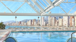 A swimming pool with a cityscape view, lounge chairs, and a glass roof structure. -  KAKTUS Hotel Benikaktus (Benidorm)