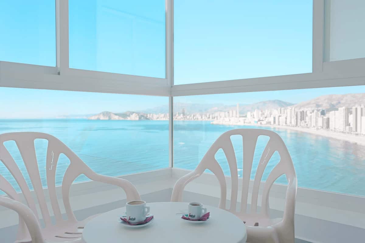 A room with a large window overlooking a coastal city and ocean. There are two white chairs and a small table with two cups of coffee.