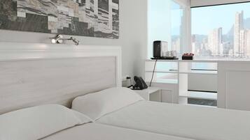 Un dormitorio moderno con vista a la ciudad, cabecera blanca, mesita de noche con teléfono y un pequeño estante con objetos decorativos.