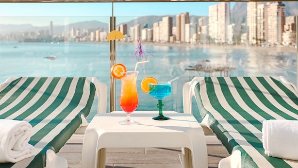 A poolside lounge with two striped lounge chairs, a table with two drinks, and a cityscape view in the background. -  KAKTUS Hotel Benikaktus (Benidorm)