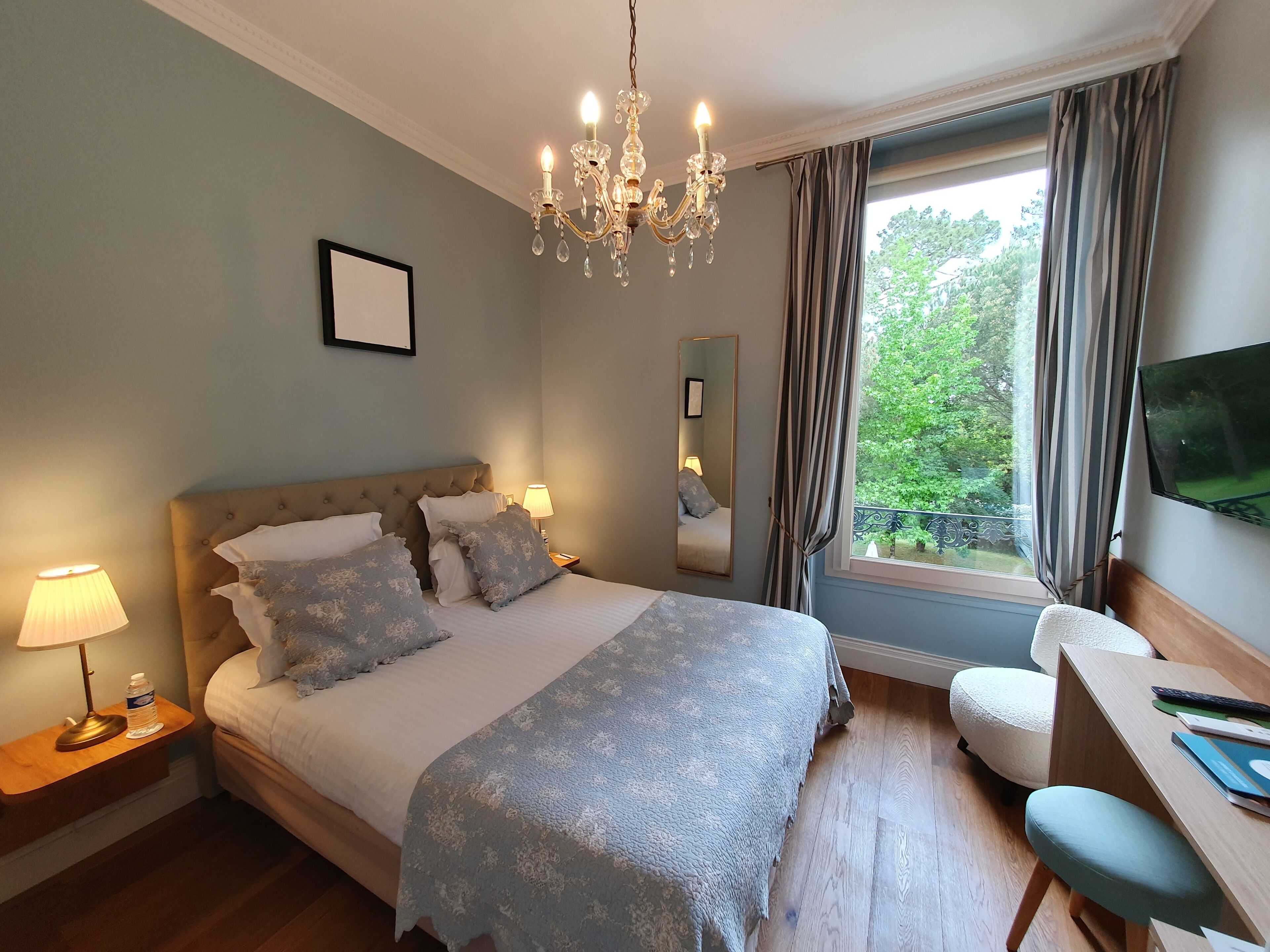 A bedroom with a large bed, bedside lamps, a chandelier, a mirror, a TV, and a view of greenery outside.