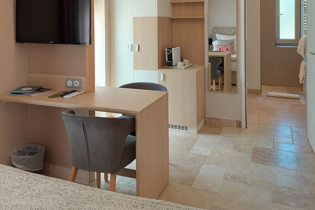 A modern room with a flat-screen TV, a desk with a chair, a wardrobe, a coffee machine, and a bed with a patterned headboard.