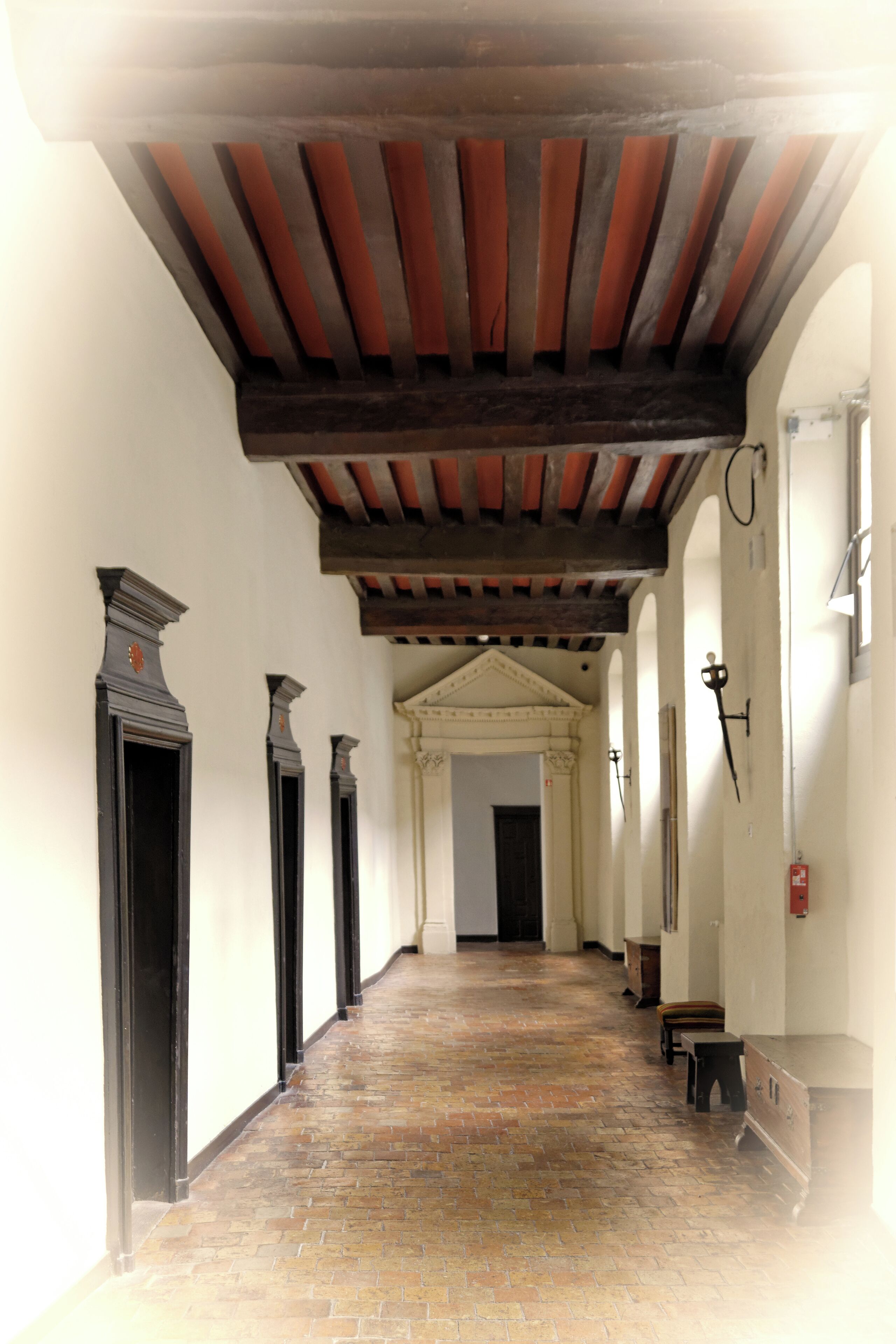 A corridor with a tiled floor, wooden beams on the ceiling, and black-framed doors.