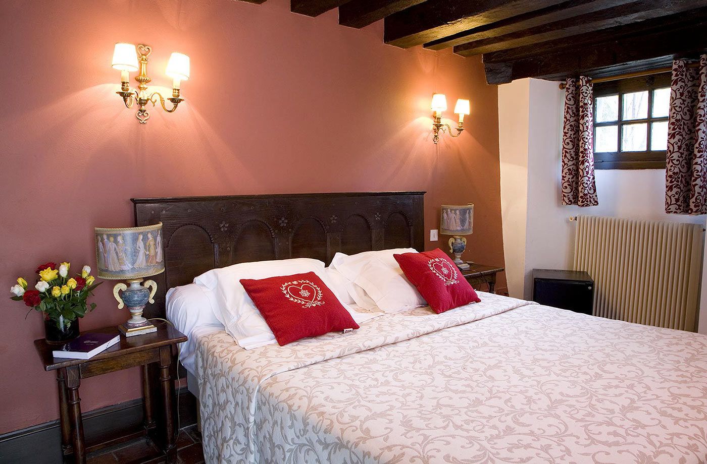 Un lit bien fait, avec des oreillers blancs et rouges, deux lampes fixées au mur, une petite table avec une lampe et un vase rempli de fleurs.
