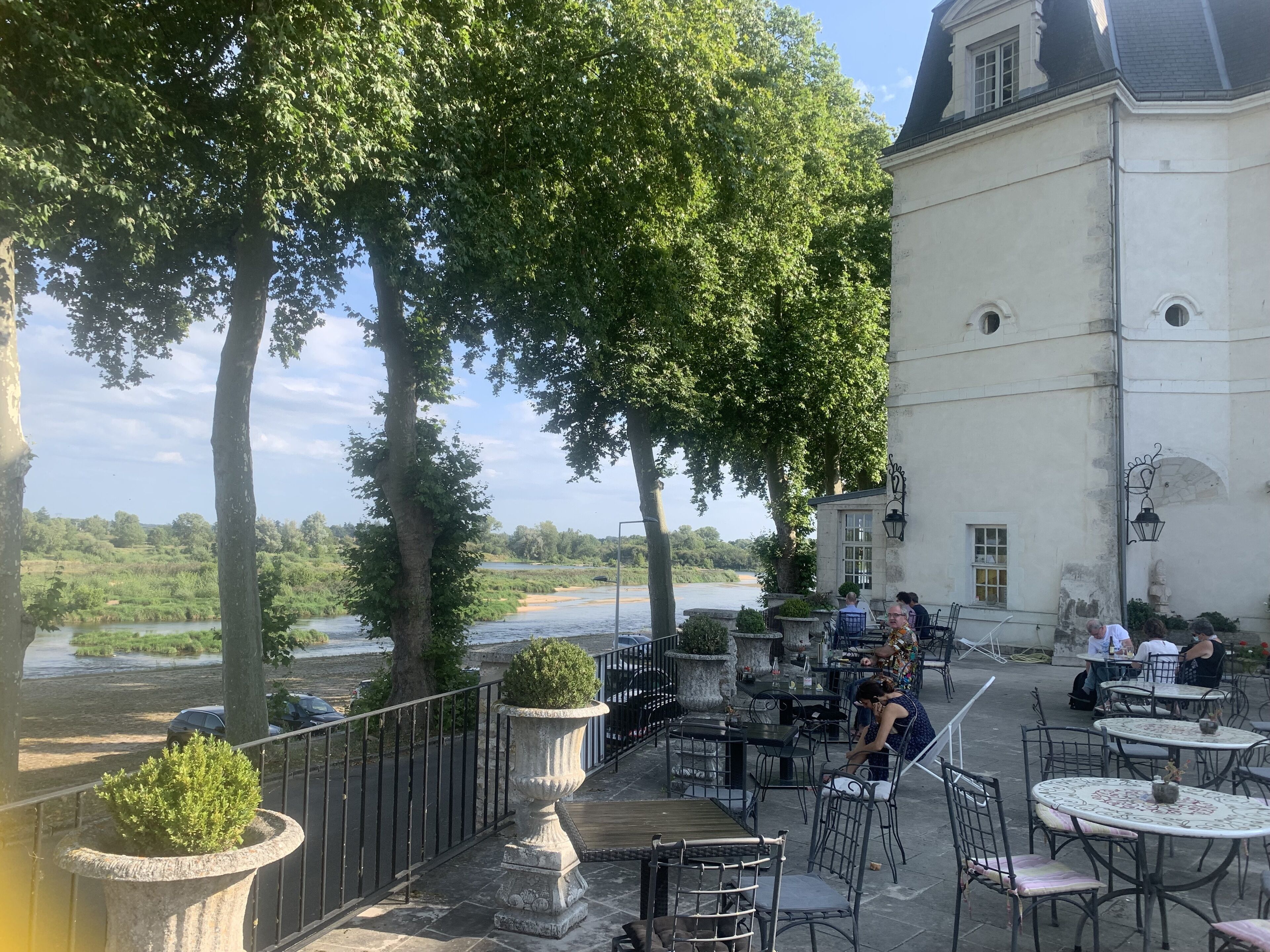 An outdoor cafe with tables and chairs, overlooking a river and surrounded by trees.