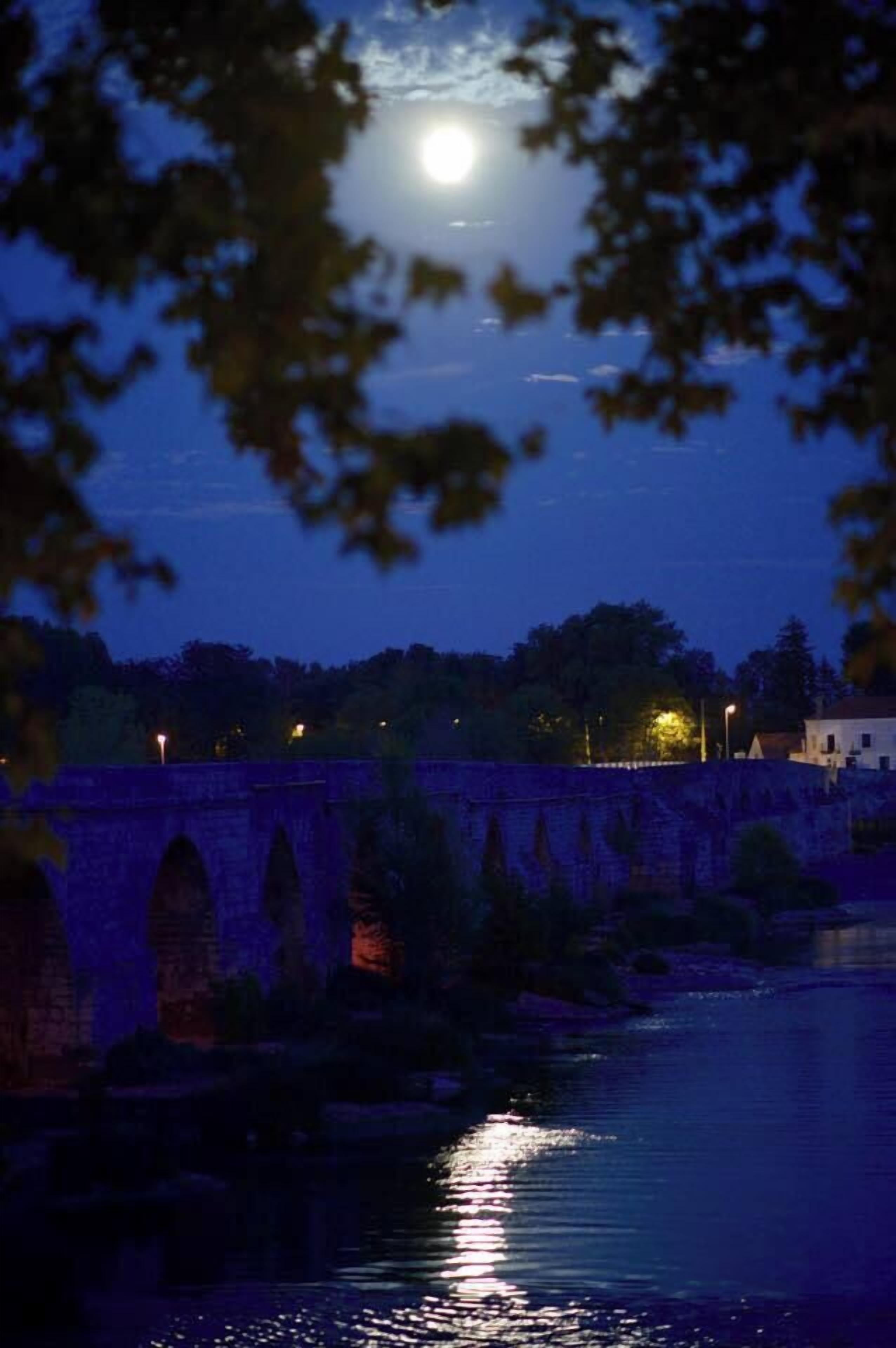 Un pont de pierre enjambant une rivière, de nuit, avec une pleine lune en arrière-plan.