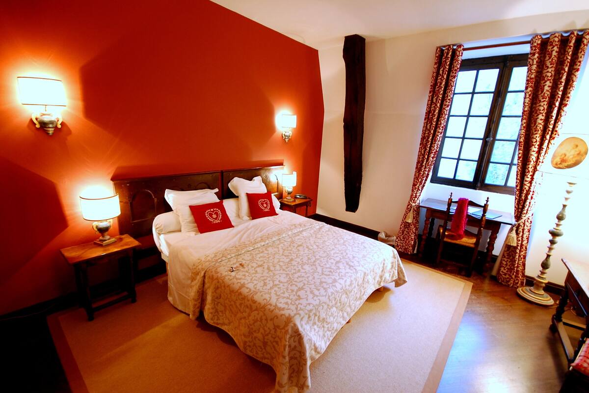 A hotel room with a large bed, two bedside tables with lamps, a window with curtains, and a small table with a chair.