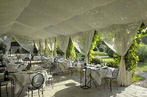 Un área de comedor al aire libre con mesas dispuestas para una comida, rodeada de telas blancas y follaje.